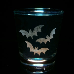 May include: A clear shot glass with a black background. The glass features three silver bat silhouettes. The bats are arranged in a triangular pattern on the front of the glass. The glass is illuminated from below.
