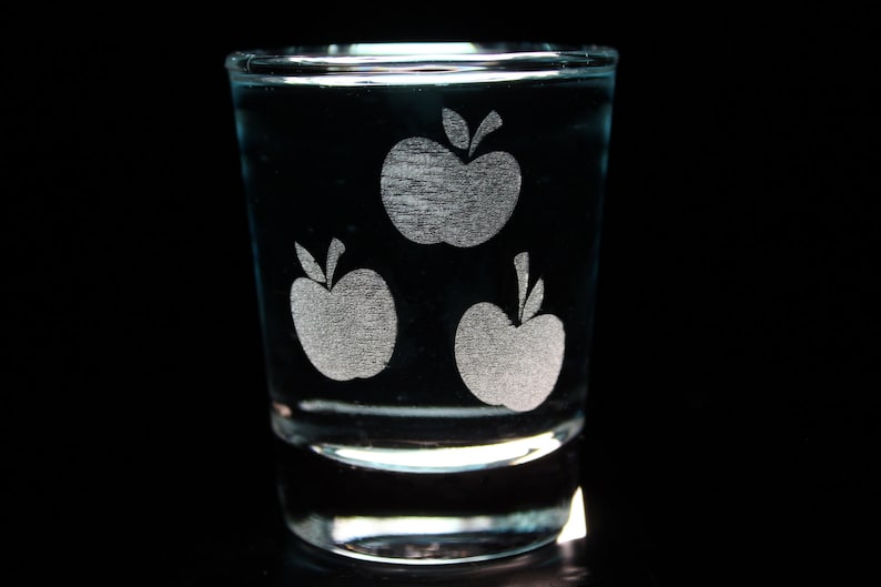 Pode incluir: Um copo de shot de vidro transparente com tr&ecirc;s desenhos de ma&ccedil;&atilde;s gravados. As ma&ccedil;&atilde;s s&atilde;o brancas e t&ecirc;m um caule e uma folha.