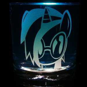 May include: A clear glass tumbler with a white etched design of a cartoon pony wearing sunglasses. The pony has a blue mane and a horn.