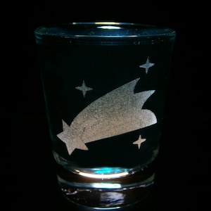 May include: A clear glass shot glass with a white etched design of a shooting star and stars. The glass is filled with a clear liquid.