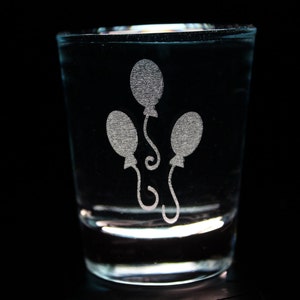May include: A clear glass shot glass with an etched design of three balloons. The balloons are white and have a simple, rounded shape.