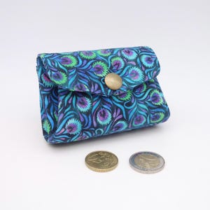 May include: A small fabric coin purse featuring a repeating pattern of stylized peacock feathers in shades of blue, purple, and green. The pouch has a triangular flap secured by a single brass-colored metal snap button, positioned near two coins.