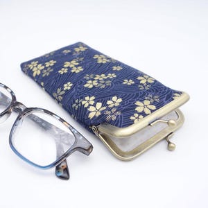 May include: Eyeglasses and a navy blue glasses case with a gold floral pattern. The case has a metal clasp. The eyeglasses have a blue and grey frame.