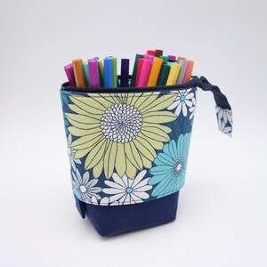 Puede incluir: Un estuche de tela azul marino con diseño telescópico, decorado con un estampado floral de margaritas grandes en verde lima, turquesa y blanco. El estuche contiene varios rotuladores y bolígrafos de colores y tiene un tirador de cremallera a juego.