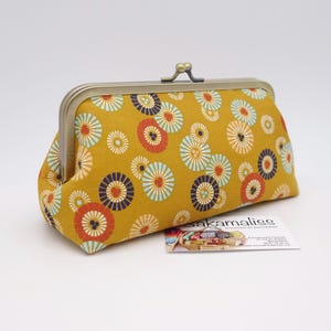 May include: A mustard yellow fabric coin purse featuring a repeating pattern of circular floral motifs in orange, teal, and dark blue. The purse has a metallic kiss-lock frame closure at the top and is positioned next to a small white business card.