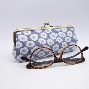 May include: A fabric eyeglass case with a purple background and a repeating pattern of white flowers with light blue petals and orange centers, featuring a bronze-toned metal kiss-lock clasp, positioned behind a pair of brown tortoiseshell glasses.