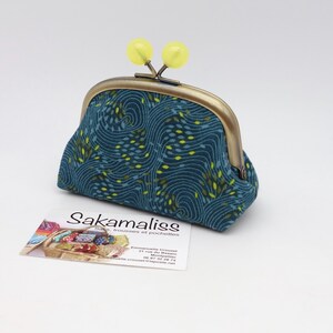 May include: A teal fabric coin purse with a dark blue swirling pattern and small yellow leaf-like accents. The purse features a bronze-toned metal kiss-lock frame topped with two round, translucent yellow beads as the clasp closure mechanism.