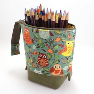 Peut inclure: Un porte-crayons en tissu vert avec une fermeture éclair et une poignée. Le porte-crayons est décoré d'un motif de hiboux et de feuilles. Le porte-crayons est rempli de crayons de couleur.