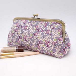 May include: A rectangular fabric clutch with a kiss-lock bronze metal frame, featuring a lavender background with a dense pattern of small white and pink cherry blossom flowers. Two makeup brushes and a small cosmetic tube lie in front.