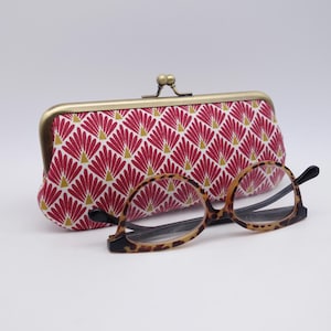 Puede incluir: Un estuche de tela para gafas con marco de latón y cierre de boquilla, con un patrón repetitivo de abanicos magenta y pequeños triángulos dorados sobre fondo blanco, acompañado de unas gafas con estampado de leopardo marrón y negro.