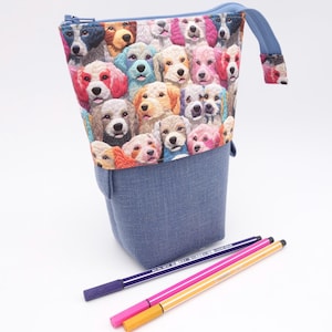 Puede incluir: Estuche de lápices de mezclilla azul con un patrón colorido de varias razas de perros. El estuche tiene cierre de cremallera y un lazo de tela. Tres bolígrafos de colores están al lado del estuche.