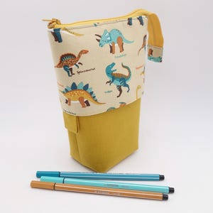 May include: A yellow pencil case with a dinosaur pattern, featuring various dinosaurs in blue, brown, and green on a cream background. Three pens in blue and brown are placed in front of the case. The case has a zipper closure.