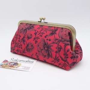May include: A rectangular fabric clutch purse featuring a vibrant red background with a dark floral pattern. The purse has a metallic brass-toned frame with a double ball clasp closure at the top. The fabric displays detailed botanical illustrations.