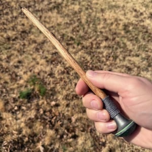May include: A handcrafted wooden wand with a light brown finish. The wand has a dark grey grip wrapped with a textured material and a dark green base. The wand is held in a hand against a blurred outdoor background.
