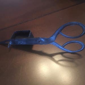 May include: A black metal candle snuffer with a square-shaped candle holder attached to the end of the handle. The snuffer is shaped like a pair of scissors.
