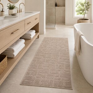 May include: A rectangular runner rug in a neutral beige tone featuring a textured geometric pattern of interlocking rectangles. The rug has a low-pile construction with finished edges, positioned on a light-colored tile floor in a bathroom.