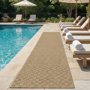 May include: A rectangular, beige outdoor rug with a geometric pattern. The rug is placed next to a swimming pool, with lounge chairs and umbrellas in the background. The rug is made of a woven material.