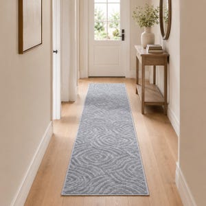 May include: A long, rectangular runner rug featuring a light gray color palette with a repeating pattern of concentric, wavy circular shapes. The rug has a low-pile texture and is positioned centrally on a light-colored wooden floor in a hallway.