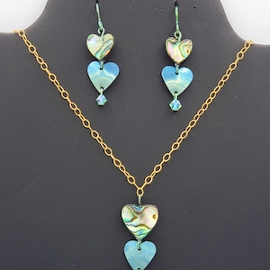 May include: A gold chain necklace with a pendant featuring three layered iridescent blue and green heart-shaped charms and a crystal accent. Matching earrings with three layered iridescent blue and green heart-shaped charms and crystal accents.