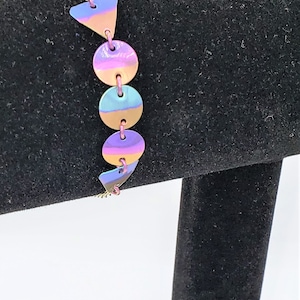 May include: A gold chain bracelet with iridescent pink, blue, and purple geometric shapes. The shapes are a triangle and circles.