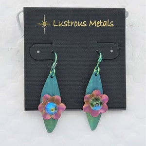 May include: A pair of teal, pink, and iridescent blue flower earrings with a teardrop shape. The earrings are on a black card with the text "Lustrous Metals".