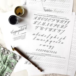 Puede incluir: Un conjunto de hojas de papel blanco con el alfabeto Copperplate en caligrafía negra, líneas de práctica e instrucciones. Se incluye una pluma caligráfica de madera marrón oscuro con punta metálica y un frasco abierto de tinta negra.