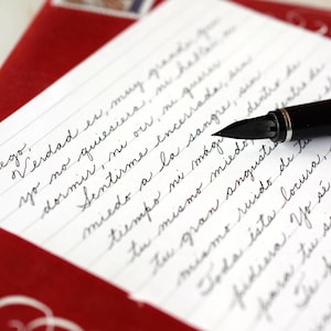 May include: A black fountain pen rests on a piece of white lined paper featuring handwritten Spanish cursive text. The paper is positioned against a deep red background, creating a contrast between the dark ink, the white page, and the red surface.