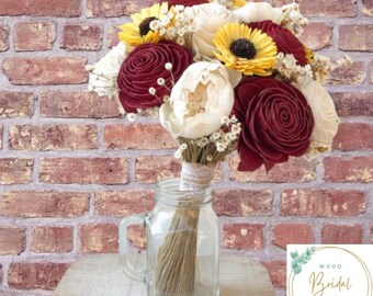 Puede incluir: Un ramo de flores artificiales con flores de madera en color burdeos oscuro, amarillo brillante y crema, mezcladas con pequeñas flores blancas de relleno. Los tallos están envueltos en encaje y colocados en una jarra de cristal.