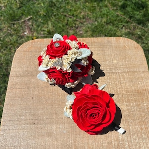 Puede incluir: Un boutonniere de rosa roja y un ramo de rosas rojas más grande con aliento de bebé blanco y acentos plateados.