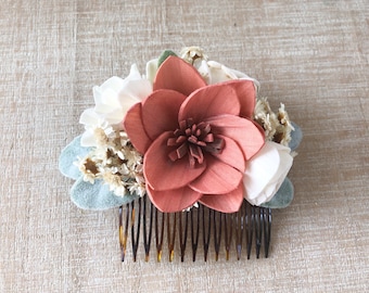 Puede incluir: Una peineta decorada con una gran flor de madera color terracota en el centro, rodeada de flores blancas más pequeñas, flores secas de relleno en tono beige y hojas de fieltro verde salvia, sobre una base de peine marrón carey.
