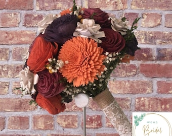 Puede incluir: Un ramo de flores artificiales en tonos naranja, rojo oscuro y crema, con una gran flor central naranja estilo dalia, rosas de color púrpura oscuro y flores de papel con notas musicales. El mango está envuelto en cordel y encaje blanco.