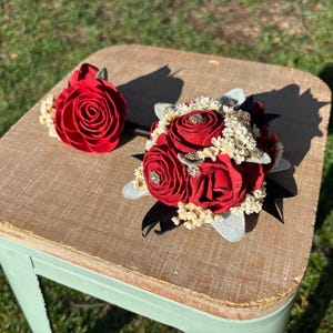 Könnte beinhalten: Ein roter und weißer Blumenstrauß mit einem einzelnen roten Rosenanstecker. Der Strauß besteht aus Seidenblumen und hat ein schwarzes Band.