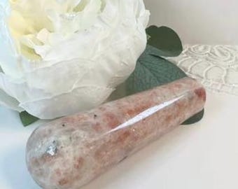 May include: A polished, cylindrical sunstone crystal wand featuring a mottled texture with shades of peach, cream, and light brown. The stone has a smooth, reflective surface and is positioned next to a large, white, layered flower.
