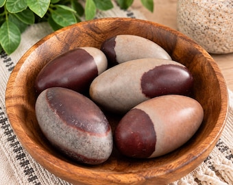 May include: A collection of four smooth, oval-shaped Shiva Lingam stones resting inside a round wooden bowl. Each stone features a distinct two-tone color pattern, blending shades of earthy brown, deep maroon, and light tan in a polished finish.
