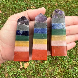 May include: Three polished crystal towers, each featuring seven horizontal bands of stacked gemstones in a spectrum of colors including purple, gray, blue, green, yellow, pink, and red, arranged in a pointed obelisk shape.