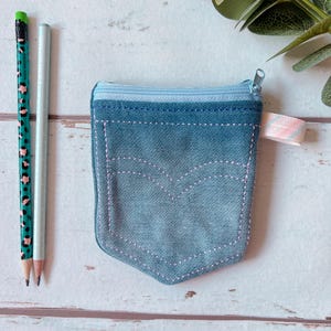 Könnte beinhalten: Eine kleine blaue Jeans-Reißverschlusstasche mit einer rosa gestickten Tasche. Die Tasche hat einen Reißverschluss und ein hellblaues Stofffutter.