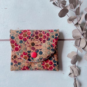 Könnte beinhalten: Eine kleine Brieftasche aus Kork mit einem roten Knopfverschluss. Die Brieftasche hat ein farbenfrohes, geometrisches Muster mit roten, blauen und grünen Akzenten.