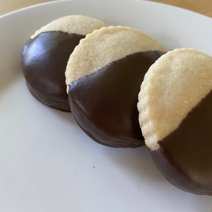 May include: Three round cookies with a half-circle of dark chocolate on top. The cookies are arranged in a row on a white plate.