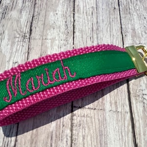 May include: A personalized keychain wristlet in pink and green. The pink fabric is accented with a green ribbon embroidered with the name "Mariah" in a cursive font. A gold-colored metal clasp and keyring complete the design.