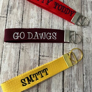 May include: Three fabric keychains in red, maroon, and yellow. The red keychain has "HOTTY TODDY" embroidered in black. The maroon keychain says "GO DAWGS" in white. The yellow keychain says "SMTTT" in black.