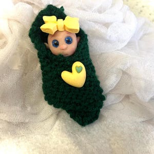 May include: A small doll with blue eyes and a yellow bow is wrapped in a green knitted blanket with a yellow heart.