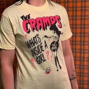 May include: Pale yellow t-shirt featuring the band name "The Cramps" in pink, with a black and white graphic of a woman and the text "What's Inside A Girl?" in black and pink. A skull graphic is also present.
