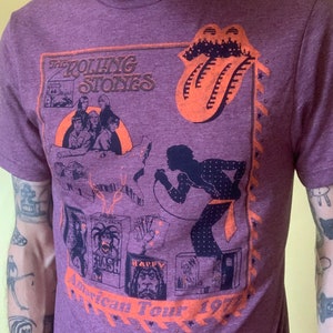 Puede incluir: Camiseta morada con un gráfico de estilo vintage de la gira americana de The Rolling Stones de 1972. El gráfico presenta una silueta de un hombre cantando en un micrófono, una banda de músicos y el icónico logotipo de la lengua de los Rolling Stones.