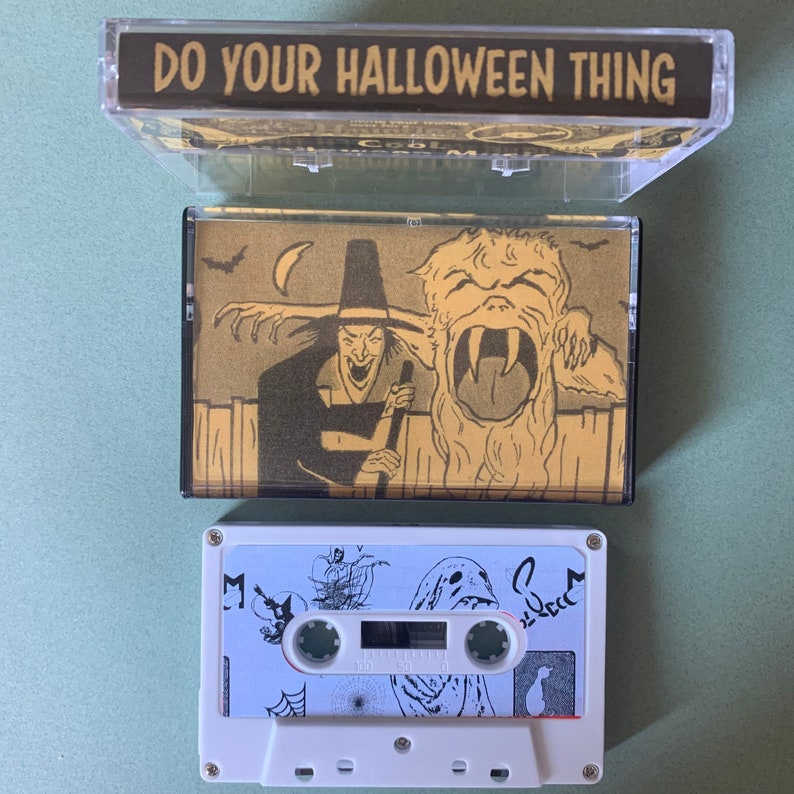Puede incluir: Una cinta de casete vintage con una caja negra y dorada. La caja tiene una etiqueta negra con el texto "DO YOUR HALLOWEEN THING". La cinta de casete tiene una etiqueta blanca con una ilustraci&oacute;n en blanco y negro de una bruja, un monstruo y un fantasma.