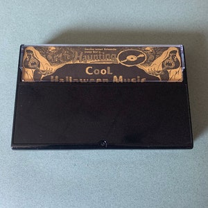 Puede incluir: Una cinta de casete negra con una etiqueta dorada que dice "Invita a tus amigos a mi Haunting Cool M&uacute;sica de Halloween". La etiqueta presenta una imagen de dibujos animados de un monstruo con un tocadiscos.