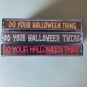 Puede incluir: Tres cintas de cassette con el texto "Do Your Halloween Thing" impreso en ellas. La cinta superior tiene texto amarillo, la cinta del medio tiene texto verde y la cinta inferior tiene texto rojo.