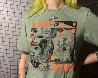 Może przedstawiać: Zielony T-shirt z czarno-białym nadrukiem graficznym czaszki i słowami "Know Fear". Grafika zawiera również różne inne symbole i wzory.