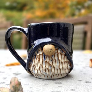 May include: A ceramic mug featuring a dark blue glazed upper section and a textured, off-white lower section with vertical brown grooves. A small, round, speckled tan nose is centered on the front, resembling a gnome character.