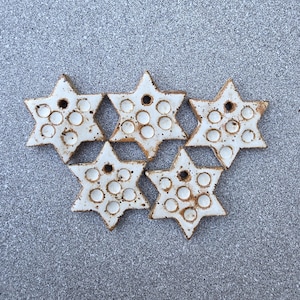 May include: Five star-shaped ceramic ornaments with a white glazed finish and brown speckled edges. Each star features a series of small circular indentations across the surface and a single hole at the top point for hanging purposes.