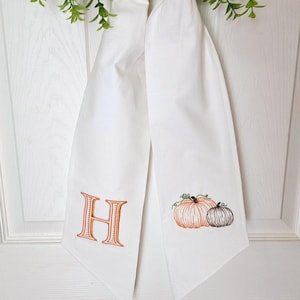 May include: White fabric door hanger with an embroidered orange letter "H" and two pumpkins. The pumpkins are orange and brown with green vines. The hanger is tied at the top with green foliage.